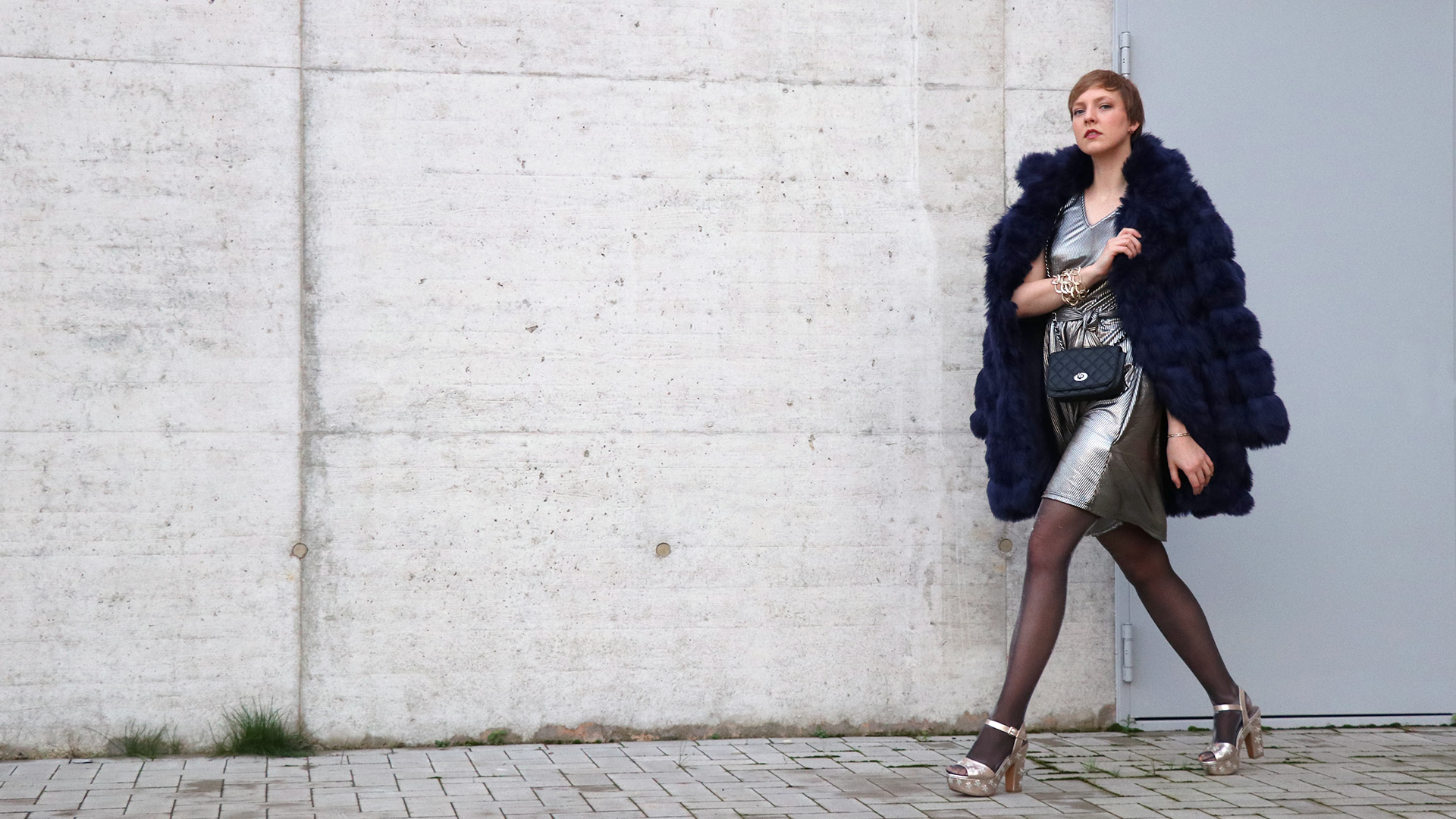 Frau mit dunkelblauem Kunstfellmantel und bronze-metallic Kleid läuft an einer Wand entlang.