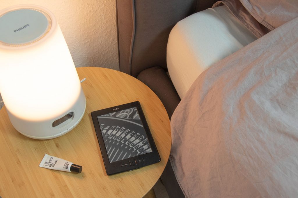 Nachttisch neben dem Bett mit einer dimmbaren Lampe, einem E-Reader und einem Lipgloss