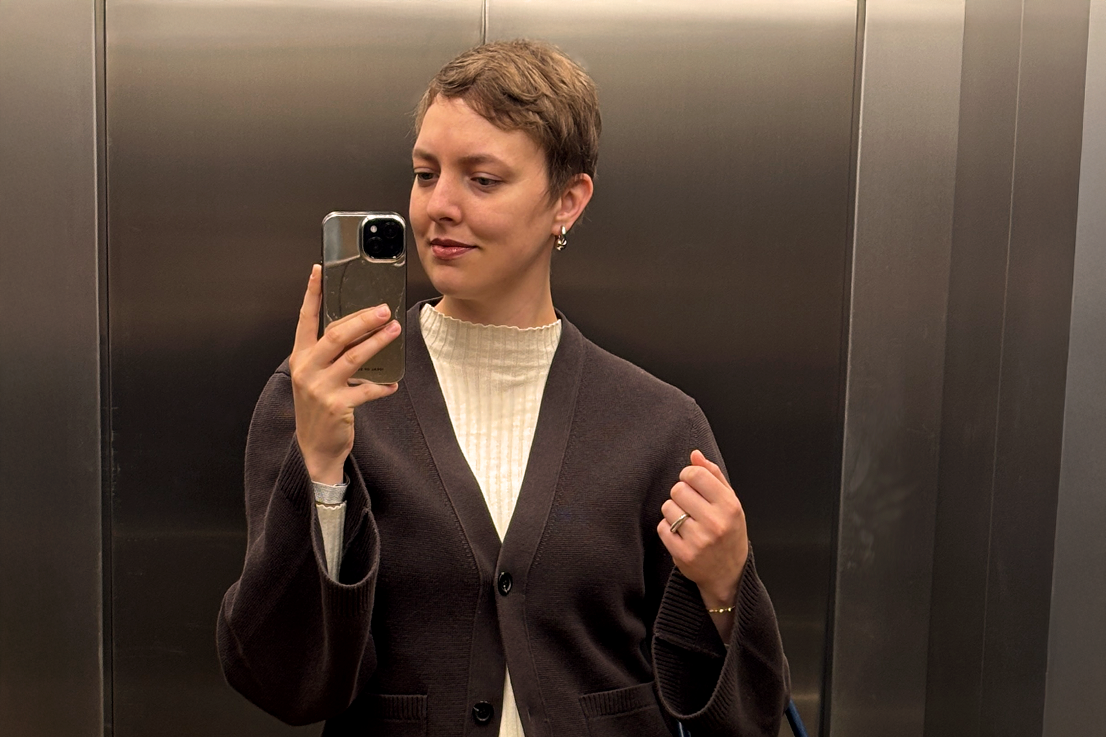 Biene von Sustylery steht im Aufzug und macht im Spiegel ein Selfie von sich in ihrem Outfit mit brauner Strickjacke und cremefarbenen Oberteil