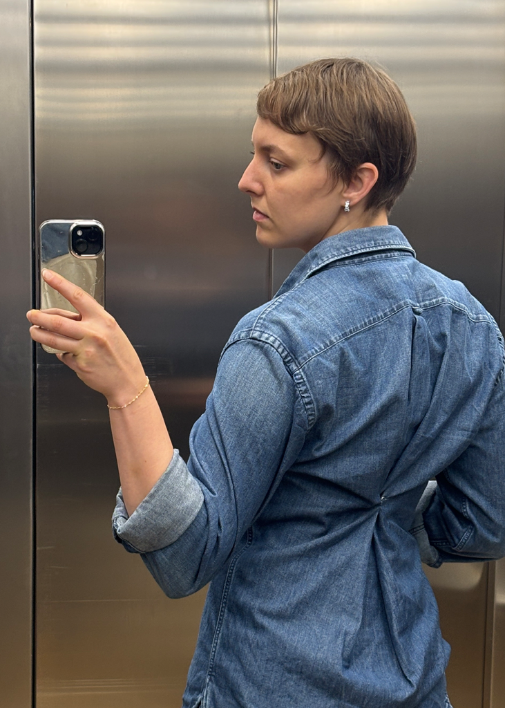 Biene von Sustylery steht im Aufzug und macht im Spiegel ein Selfie von sich in ihrem Outfit mit Jeanshemd, das sie im Rücken mit einer Sicherheitsnadel zusammengeheftet hat
